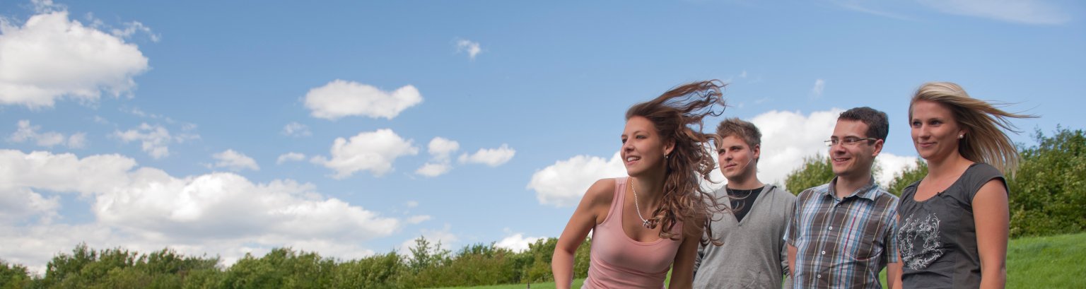 Eine Gruppe junger Leute steht am  Ufer eines halb zu sehenden Gewässers im leichten Wind, der die Haare fliegen lässt.