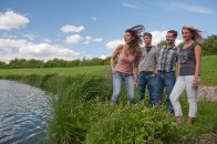 Eine Gruppe junger Leute steht am Ufer eines halb zu sehenden Gewässers im leichten Wind, der die Haare fliegen lässt.